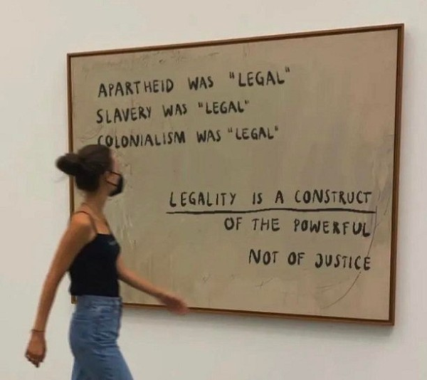 White board written with:

"Apartheid was 'legal'
Slavery was 'legal'
Colonialism was 'legal'
    LEGALITY IS A CONSTRUCT
    OF THE POWERFUL
    NOT OF JUSTICE"