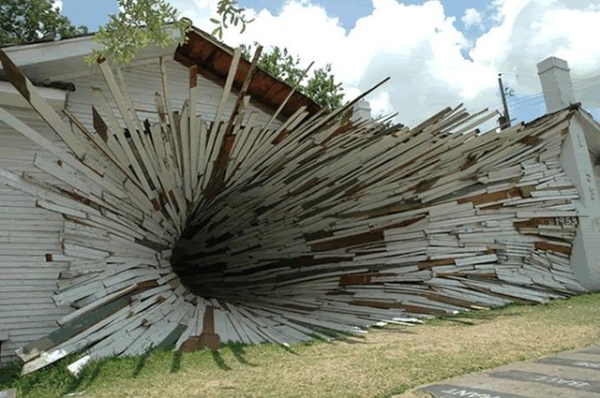 A white building with one side of it seemingly being sucked into itself. The sky is blue and full of fluffy clouds, and the trees have green leaves, but the building looks like it is being eaten by a black hole.