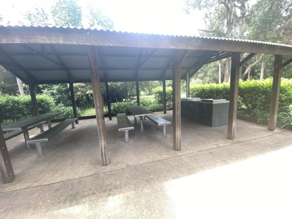 Photo of a metal roofed shelter shading metal picnic tables and benches plus some publicly available bbqs (grills).   