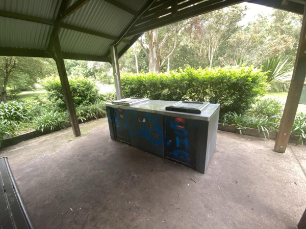 Photo of the builtin metal bbqs in the shade area. Bit of graffiti painted on the front in blue.  May be some kind of tagging but it's not very good.