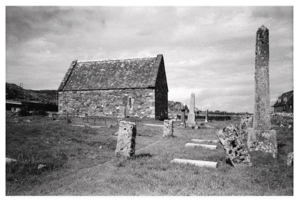 Grainy black and white film photograph showing an old stone chapel seen from within a graveyard.