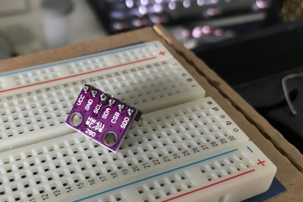 A BME280 temperature, pressure and humidity sensor placed on a breadboard. In the background there's a bokehed keayboard and a desk.