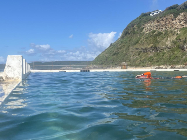 Photo of the blue green water at the lap lane end of the pool with a freestyle swimmer in a bright orange and black swimming outfit (with sleeves and leggings - so could be wetsuit or rashie and legging combo). The swimmer's left arm is raised mid stroke and one of their legs has kicked up almost to the surface. Blue sky, clouds and overgrown rocky hillside in the background.