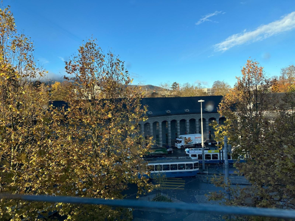 A view from an upper window, overlooking autumn trees in the foreground, an old tram below with a wide curved station behind and deep blue skies above