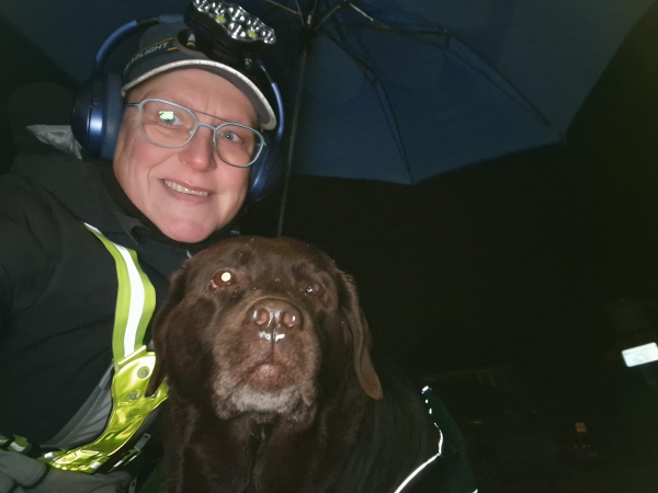 Night time selfie of a person and a chocolate brown Labrador with greying snout, both under a blue umbrella, looking at the camera. 