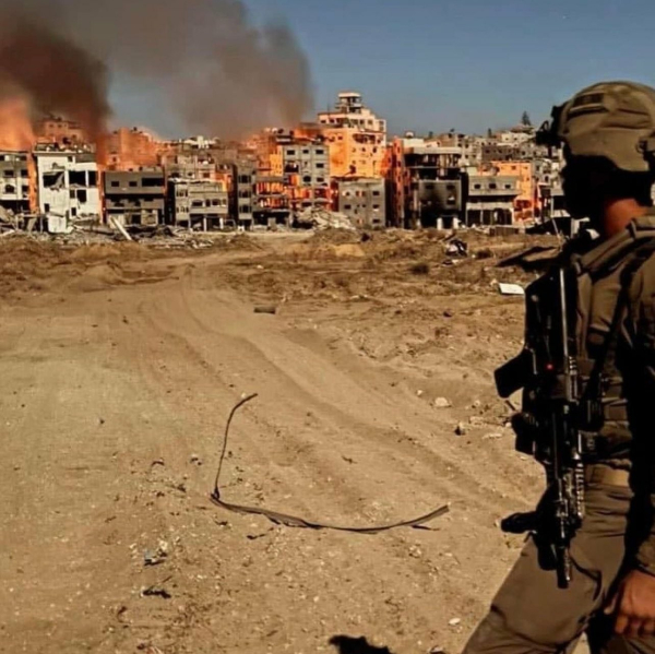 Ein israelischer Soldat steht seitlich zur Kamera, die Waffe umgehängt, er schaut zurück zur Stadt, die komplett in Brand steht. 