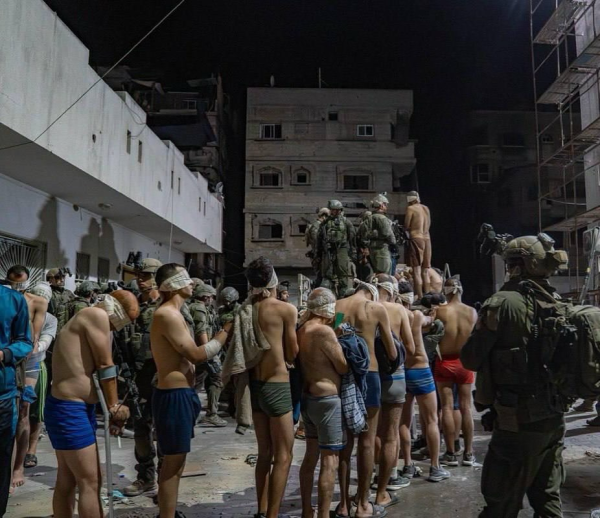 Ein Bild vom Kamal Adwan Krankenhaus im Oktober. 

Es ist Nacht bzw Dunkel,  ringsum Gebäude, teilweise mit deutliche Schäden.  In der Mitte ist ein Truck auf dem israelische Soldaten stehen, die Gefangene geladen werden. Im Bild verstreut stehen  hochgerüstete israelische Soldaten.  In einer Reihe stehen zahlreiche männliche Personen, jeden Alters.  Alle sind bis auf die Unterhose nackt, alle wurden die Augen verbunden, fast allen wurden die Hände gefesselt.  Viele von den gefangen genommen sind Healthcare Worker und Patienten, bei einigen sieht man medizinische Equipement (Zu- und Ableitungen, Sonden, Gehilfen)  