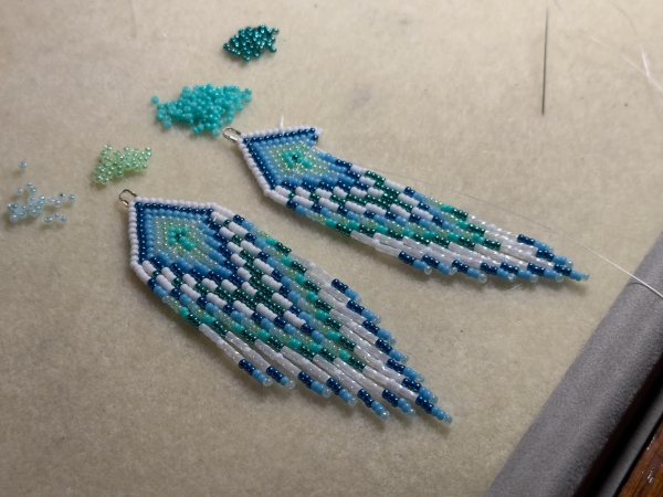 A pair of white, blue, and turquoise beaded fringe earrings in progress on a cream colored bead mat