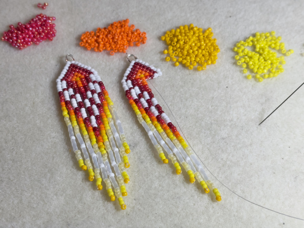 A pair of red, orange, yellow, and white beaded fringe earrings on a bead mat, one is finished and one in progress. Above the earrings are piles of red, orange, and yellow beads.