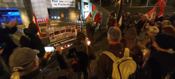 Rassemblement nocturne. Une personne parle dans un mégaphone devant des banderoles et des bougies. 