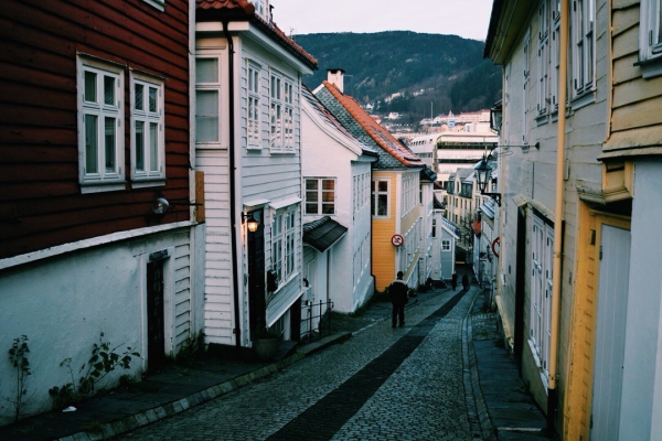 A narrow, cobblestone street lined with colorful houses. People are walking down the street, with hills and buildings visible in the background.