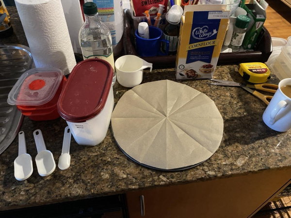 Photo of assorted pavlova ingredients and baking paraphernalia on the kitchen benchtop. There's a container for the egg whites to warm up to room temp and another for caster sugar, white vinegar bottle, cornflour packet, 3 white plastic measuring spoons, a white plastic measuring cup, and a springform tin base covered in a round of baking paper.