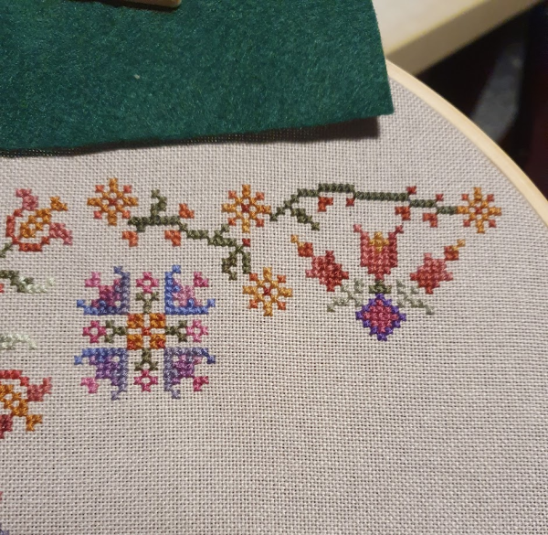 Cross stitch on taupe coloured fabric. Along the top is a small green vine with orange coloured flowers, below that are two separate flowers - one in shades of purple and the other mostly red and pinks. 