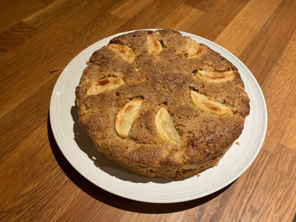 Photo of an apple cake on a white plate, on an oak worktop. The cake has a few slices of apple arranged on the top (more diced in the sponge) and a crunchy demerara sugar topping.