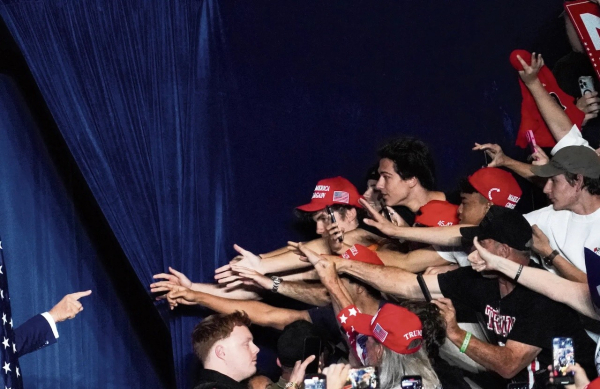 A photo of Donald Trump, only visible at the left edge by way of his hand pointing, from behind a sliver of an American flag, at a group of his supporters, almost all male and clad in MAGA gear, stretching their hands out towards  him from what is probably some sort of tiered seating. If they aren’t holding phones that is.

The background is something like a dark blue velvet, bunched up in a way that creates an almost perpendicular line to the angle created, from the bottom left to the top right, of Trump’s pointing hand, and the rising ‘wave’ of his supporters.

It presents an image that feels similar to Michelangelo’s fresco, ‘The Creation of Adam’, except for the fact that it is young white men both creating Trump and wanting to be created by him in reverse.