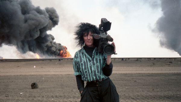 A still from the film Never Look Away. A white woman with unruly black hair, black clothing and a blue striped shirt stands in a desert. She is holding a large camera on her shoulder and smiling as a fire burns on the horizon, thick black smoke billowing up into the pale sky.