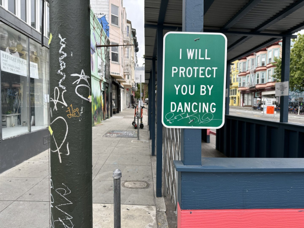 Photo of a sign attached to the vertical, wooden post of a dining “parklet” shelter on a city street and sidewalk. The scene is empty of people, and the natural light is gray and overcast. The overall effect is lonely.

The sign itself is green and white and resembles a typical, official street parking regulation sign. It reads:

I WILL PROTECT YOU BY DANCING