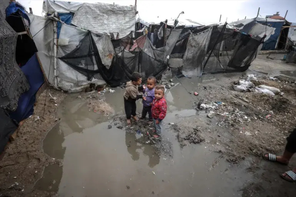 Three small boys stand on rubble midst a flooded area, surrounded by dilapidated, torn makeshift shelters.