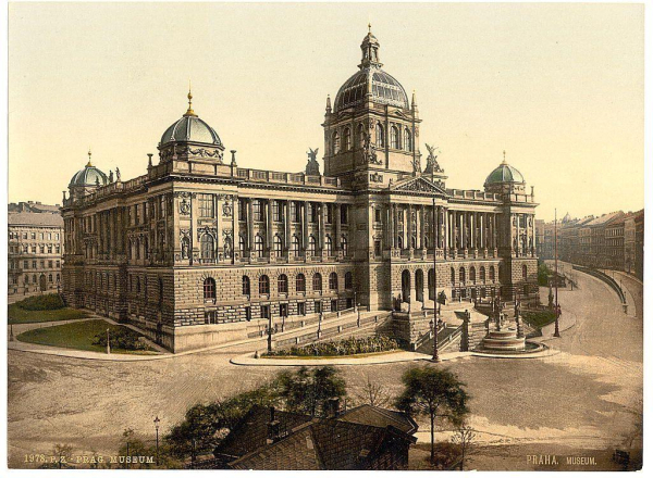 This image depicts a historic building with Baroque architectural elements, showcasing ornate details such as statues and decorative moldings. The structure has multiple stories, symmetrically arranged windows, and two prominent domed towers at the center, which are likely to be significant features of its design.

The photograph is an early 20th-century postcard or reproduction print that captures a broad view of the building from across the street. It appears aged with sepia tones, suggesting it may have been printed on photographic paper typical of the era.

In front of the museum-like structure is a spacious courtyard area with several trees and pathways leading up to its entrance. There are also some smaller buildings visible in the background along what seems like an urban street lined with lampposts or other similar structures.

The image has text at the bottom indicating "Praha, Museum," which likely refers to Prague's National Gallery museum building located on Celetná Street and known as Černíček. The exact date of this photograph is unknown but could be around 1890-1900 based on stylistic details.

The image provides a glimpse into the architectural beauty and urban planning of late Austro-Hungarian Empire's Prague, reflecting its historical significance during that time period.