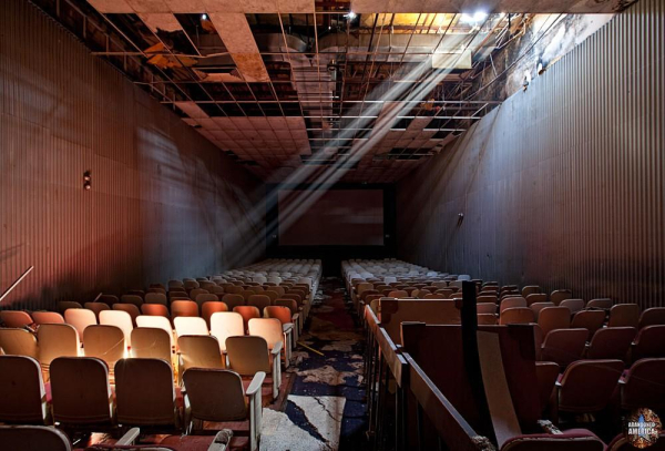 A long theater auditorium with rows of seats on either side. A shaft of light from a hole in the ceiling lances downward diagonally onto the seats.