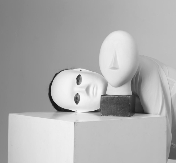 A posing box supports a white stone head of rather blank feature, on a small dark wood block base of it's own. We call it Yorick. Behind, a model lays her masked head on the box, hidden face turned toward us. Her eyes are visible.