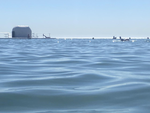 Blue ocean pool and blue sky. Small figures bathe in the sun - some in the pool, some on the edge of the pool. One sunbather is lying on their back with their feet up on the railing next to the white rounded pump house. Hot day, already 30C at 9am.