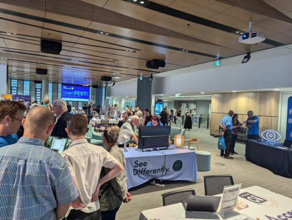 A room full of exhibitors and attendees at See Differently's Tech Fest in Adelaide.  A screen at the back of the room has the event name, and a table near our view has See Differently's banner.  The table closest to us has the NV Access logo, some fliers, a laptop and some chairs.