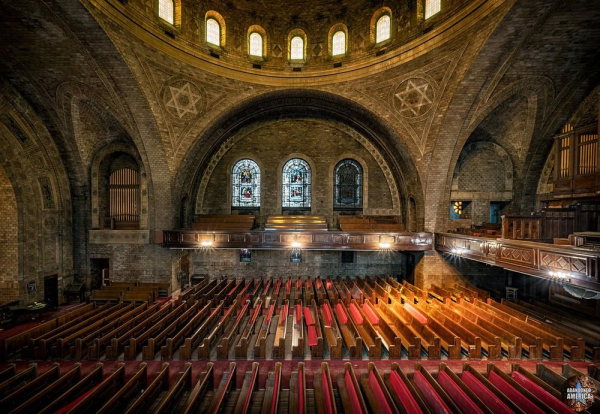 Side view of a massive house of worship, with a large arch over a balcony ahead and dozens of rows of pews below. Another balcony and organ are visible on the right.