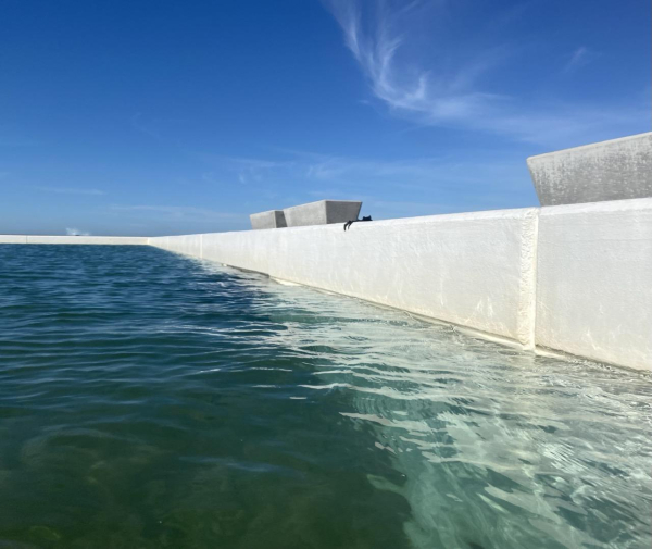 Photo of the water along the wall and southeastern corner of the main pool at the Ocean Baths. The water colour is a translucent green-blue along the side of the white painted wall, switching to a deeper blue-green where the side meets the floor of the pool. Above the white side of the pool, a matching pair of solid concrete benches are  visible, with a partial bench visible on the far right. Between the blue sky lightly feathered with cloud, the white walk and the water, the benches seem more sculptural, like an art installation, rather than simple benches.