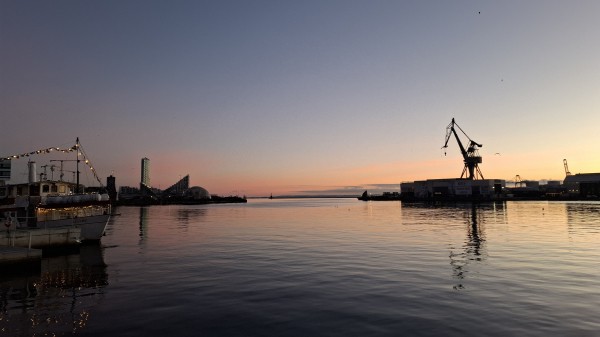 Water’s edge, mid-city Aarhus, not long after 8am today, shortly before sunrise. The sky is grey-blue, cloudless. The docks and piers across the harbour are dark, almost silhouettes with tall and low buildings in striking shapes – a huge angular crane; a narrow oblong skyscraper; a triangular apartment block; a geodome. Behind them, a stripe of deep pink and orange across the horizon. The high-tide sea is bulging with dark blue ripples, almost black, with orange and pink reflected on the surface. Just to the left, a white-and-red trawler with bunting and white Christmas lights that twinkle in reflections on the water. This is not, I think, the view you imagine when you hear the word ‘commute’.