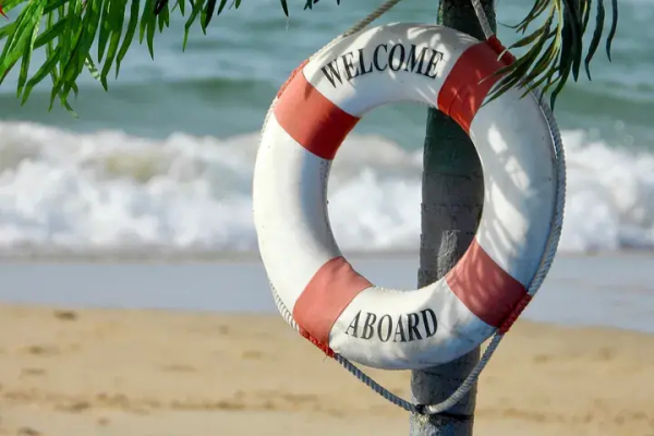 Photo of a life-saving ring, hanging on a tree near a beach with waves in the background. Printed on the lifesaver are the words Welcome Aboard.