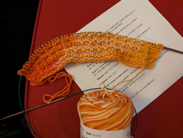 An in-progress knitted washcloth still on the circular needles, in the same orange-white cotton yarn. It's a simple pattern, a bit of a basket-weave done with knits and pearls with a seed stitch border. Only about 2-inches of it is knitted so far.