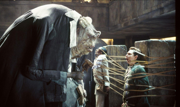 A still from the feature film adaptation of The Hitch-hiker's Guide to the Galaxy. A large, gnarled green alien wearing a tattered suit looms over two humans lashed to concrete slabs. The alien is holding a vintage microphone.