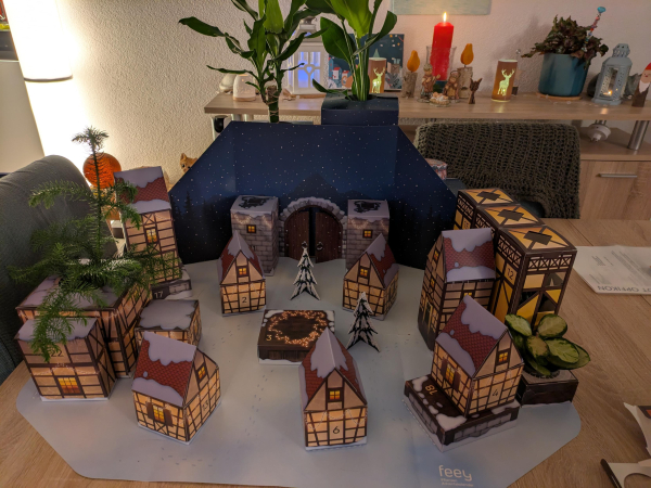 Various cardboard houses scattered village style on a table with a couple of plants poking out of some of them. Random Christmas decorations in the background on a sideboard.