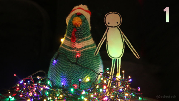 A photo of a teal and cream knitted chicken wearing a Santa hat, next to a simple figure drawn in sharpie on some white card, nestled in a mass of multi-coloured fairy lights against a black backdrop. The number one is in the top right.