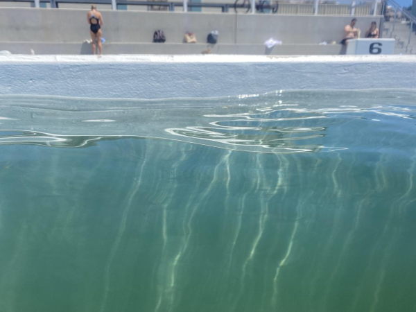 Light turquoise water streaked with long squiggly lines of light like jellyfish tentacles. Taken while swimming next to the white painted concrete wall of the Ocean Baths. In the background, a grey concrete wall with a bench that runs along it. People have left their gear on it. 2 ppl in bathers are sitting to the right; another is standing facing away from the camera to the left.