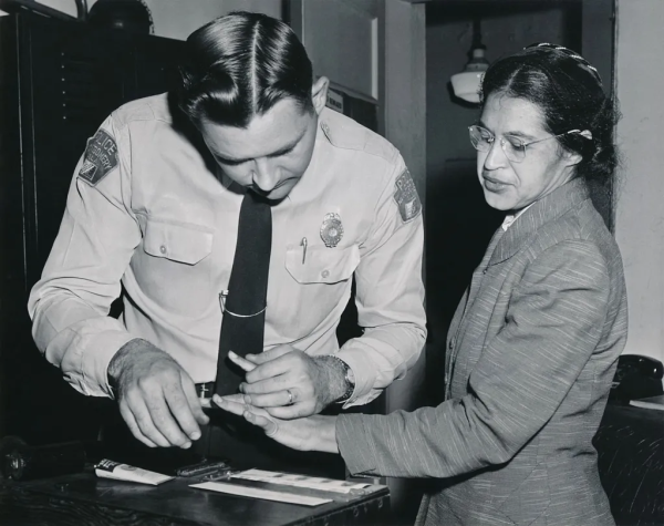 A historical photo of Rosa Parks, a Black woman wearing a suit, getting her fingerprints taken by a young white male cop.