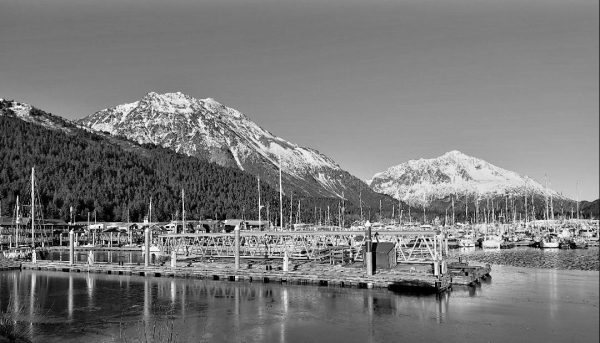 B&w photo of snow capped mountains and harbor with boats.