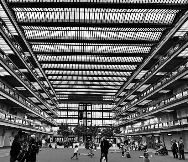 A spacious indoor area with multiple stories, featuring large glass windows and a slatted ceiling. Numerous people, including children, are scattered throughout the space, some engaging in play, and there are a few trees and greenery visible. The image is in black and white. 