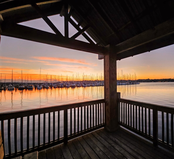 From inside a covered, wooden observation deck at the end of a pier, overlooking an approaching sunrise just beyond a busy marina lined with tied up sailboats, empty masts raised high. Light misty clouds reflect shades of pink while dark shades of orange and yellow climb from the distant horizon beyond the marina, yet illuminating the sailboats and masts over the dark waters, all reflecting incredible shadows and reflections upon the water below.