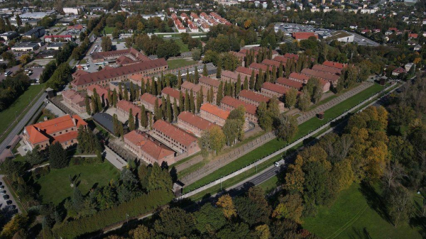 This image shows an aerial view of a large, historical complex with rows of red-roofed brick buildings, the former Auschwitz I camp - today part of the Auschwitz Memorial. The buildings are arranged in a grid pattern, surrounded by trees, greenery, and perimeter fences. There is a road on one side of the complex, with a variety of buildings and infrastructure visible in the surrounding area. The image captures the stark layout and symmetry of the camp, juxtaposed against the natural landscape and modern town buildings in the distance, conveying both historical context and present-day surroundings.