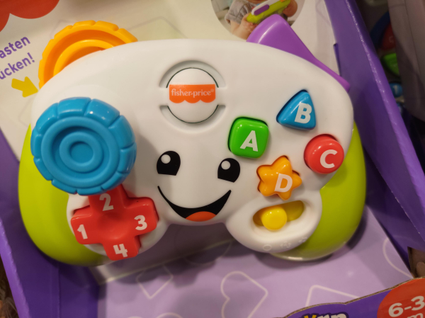 A white and green fidget toy clearly marketed towards the youngest among us, in a shape of a rounded gamepad with vastly exaggerated, colorful buttons in various shapes, a slider and a single, enormous analog on the front. The top side of the "gamepad" features a huge switch and similarily sized clicker-wheel. The device is positioned vertically in a purple box with white heart shapes on the bottom. The back side is white.