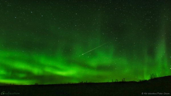 A strong auroral display beginning to break up, creating a swirling mass of green loops and spikes in the lower two-thirds of the shot. Above them, the stars are visible, in particular Ursa Major the Plough (Big Dipper) in the top left. The foreground is a silhouette of gentle hills, some trees are visible on the skyline which give scale. Near the centre of the shot, travelling from upper right to lower left, a sputtering fireball is streaking through.