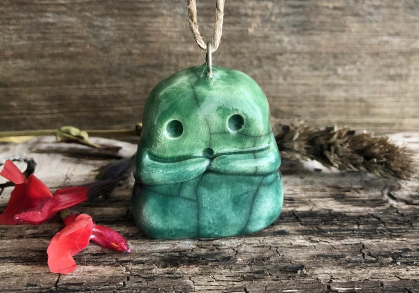 against a background of brown driftwood, a small, forest green raku sculpture of a kodama kami spirit. it has two wide, round eyes and a little round mouth, and two little arms across its front. it also has a metal loop in the top of its head and a length of twine threaded through that.
there are some red autumn-sage flowers next to it on the left, and there's a sprig of brown dried hyssop behind it on the right. the scene feels solsticey / christmasy to me, although the kodama looks a little confused by the whole thing.