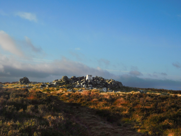 Colour photograph of a rough circle of stones in the middle of which is a white painted trig point. There is rough grass in the foreground and the sky is blue with some low whitish grey cloud.