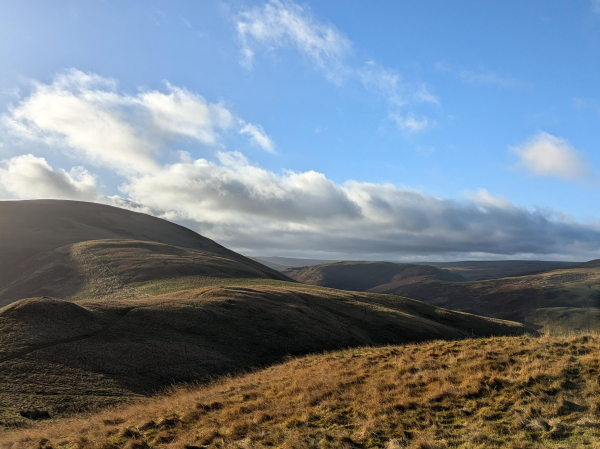 Colour photograph of a stretch of rough moorland grass with hills and valleys in the background. The low light is strong causing long deep shadows. The sky is blue with a ridge of white cloud running across close to the horizon.