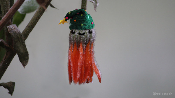 A photo of a closed orange flower, dangling down from its stem. The plant flowered valiantly through autumn, but is now covered in ice on a frosty morning. The whole thing looked quite sombre, and there's quite enough of that around, so I added a woolly Christmas tree hat (dark green, multicoloured dots for baubles, yellow start on top) and a smiling face. Everything is better with a silly hat.