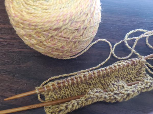 Beginning of a swatch of mottled yellow yarn, 22 stitches on US5 DPs, cake of yarn to the rear