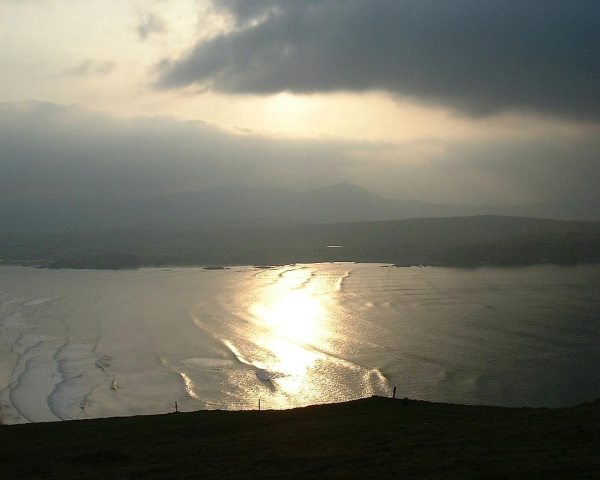 Low resolution photo, on a cliff edge. Little detail, but fencing is in the foreground, a calm sea beyond. The setting sun is reflected off the water, across to the other side of the bay. Low hills can be seen in the distance, the sun in the sky is covered by light grey cloud.