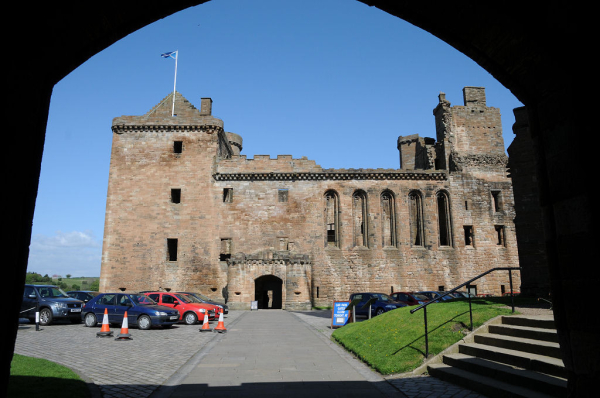 The image shows the front of Linlithgow Palace seen through the arch of the entrance gateway. The arch cuts off the sides of the frame while the palace is a large ruin made of honey-coloured stone with a tower house at its left-hand end. There are cars parked this side of it and a set of stone stesp leads up to the right beyond the arch. The sky is blue.